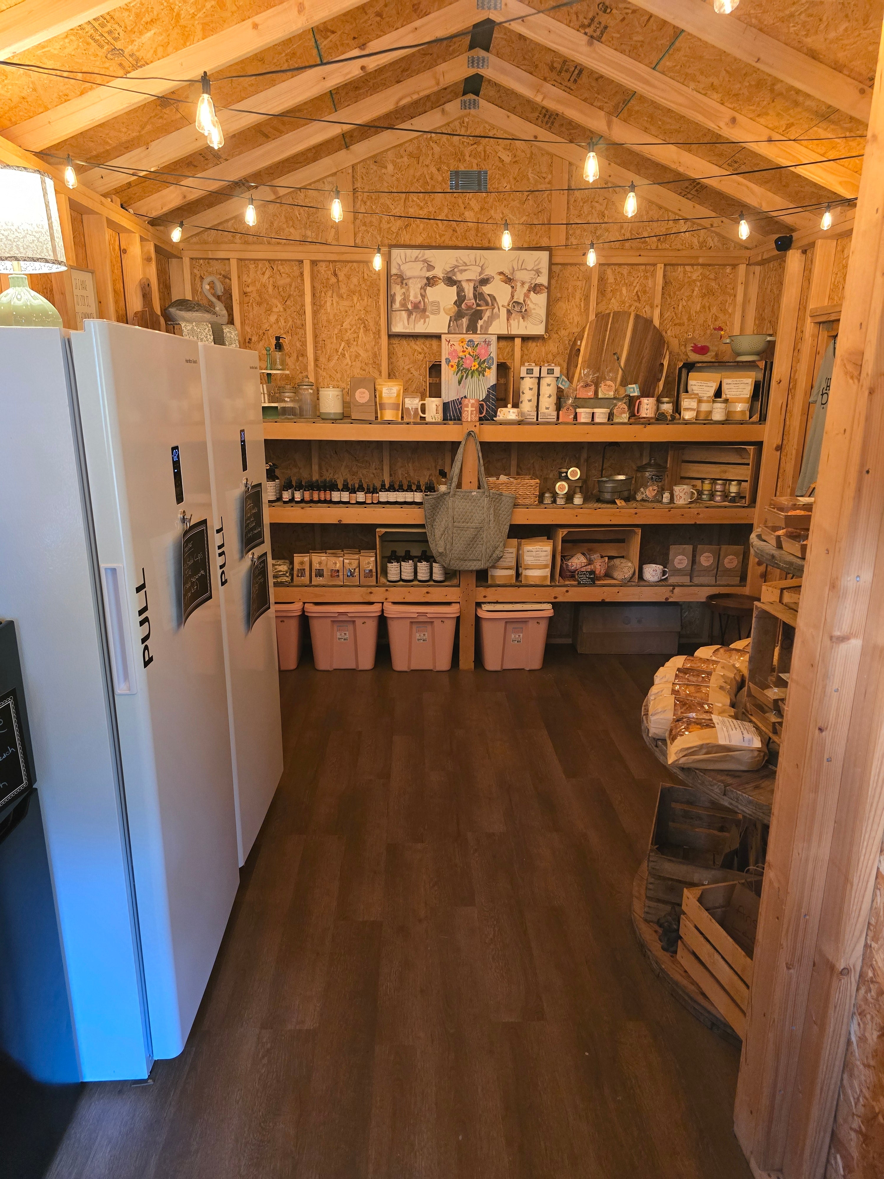 A rustic wooden shed or storage room with a high wood-beamed ceiling, warm string lights, and wood flooring. On the left is a large white refrigerator, and the center and background are filled with shelves holding jars, boxes, and home goods.